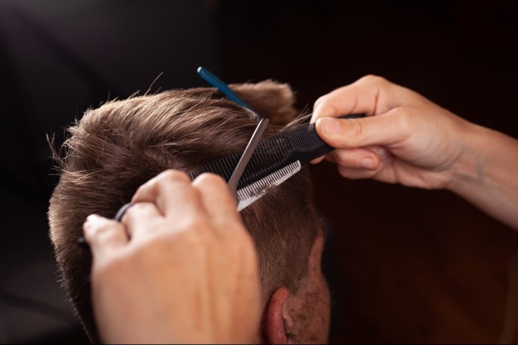 Close-up of a stylist using scissors over a comb to trim the top of a man's hair at a Barber Shop.