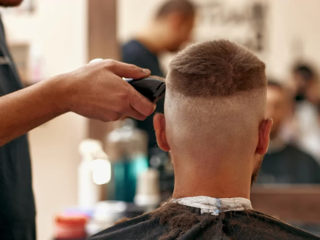 A barber expertly uses electric clippers to create a high fade haircut on a man's head in a bustling, affordable barber shop environment, showcasing services from cheap barber shops.