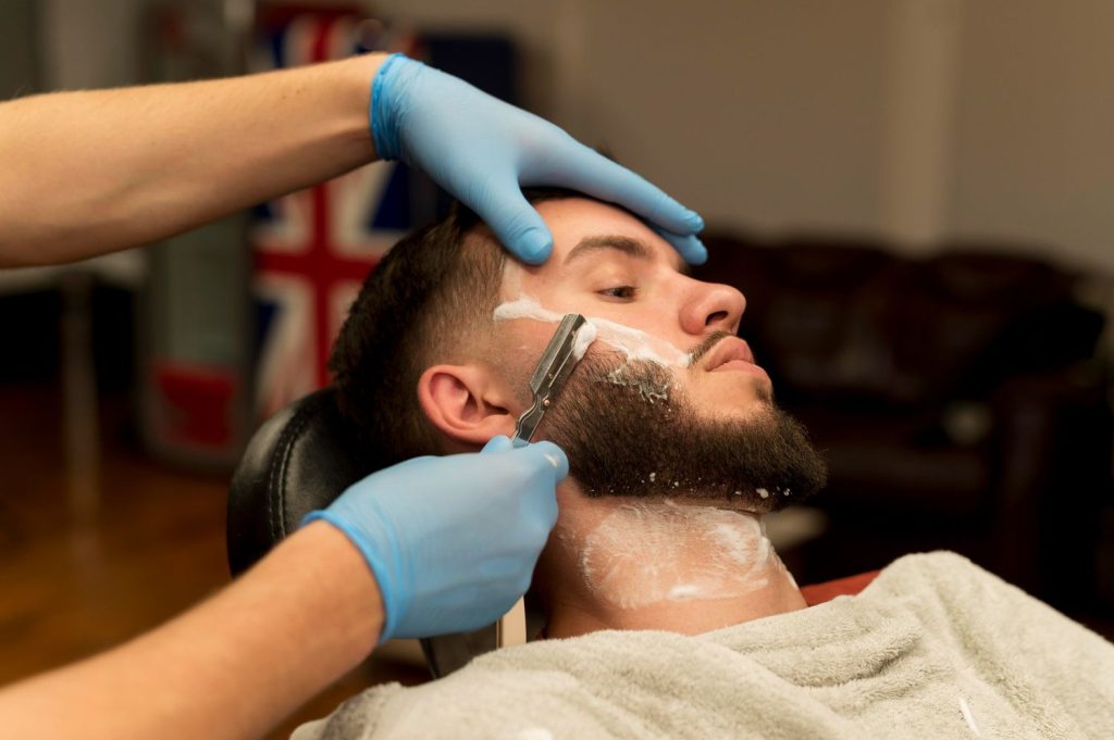 A barber shaving a man's face with a razor, covered in shaving cream, in a barbershop with a haircut focus.