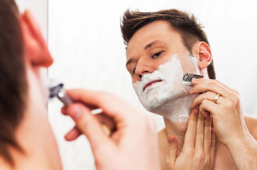 Man shaving with a razor in front of a mirror, preparing for a fresh haircut in a clean bathroom setting.