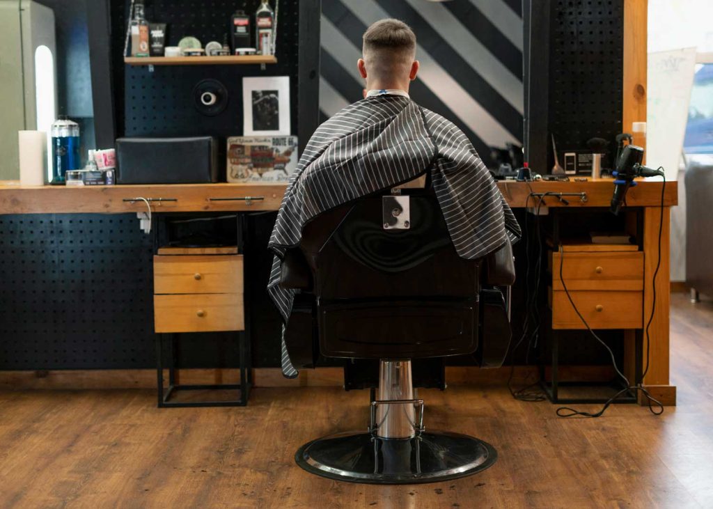 Back view of a client in a striped cape seated in a walk-in barber shop chair during a haircut, surrounded by modern tools and stylish decor for quick styling services.