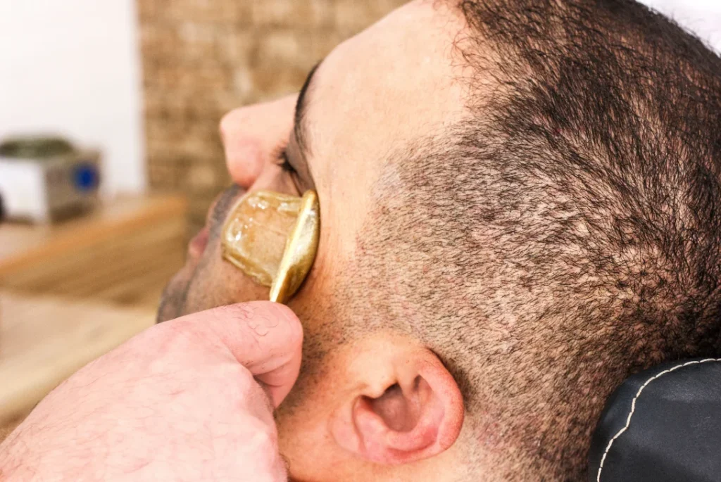 Esthetician applying green wax to a man's eyebrow in a close-up, demonstrating precise Mens Waxing.